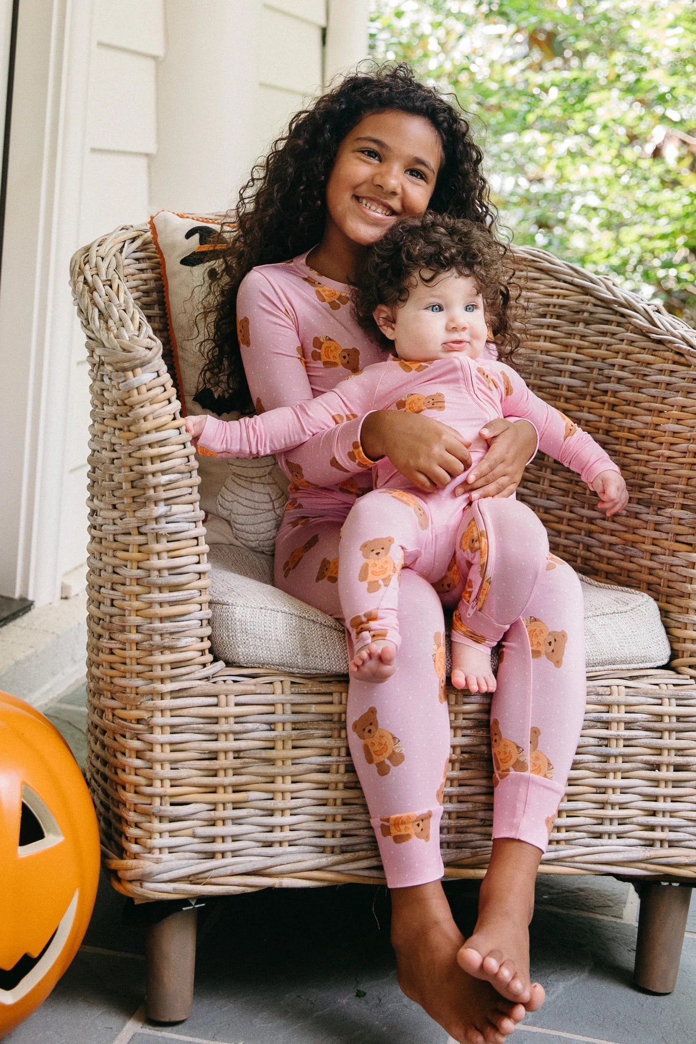 A child and a baby wearing matching pink long-sleeve pajamas with a teddy bear print, sitting together in a wicker armchair on a porch.