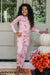 A young girl with long curly dark hair stands barefoot on a porch wearing a light pink long-sleeve pajama set with an all-over teddy bear print. She is posing next to a stacked white pumpkin and a pot of orange marigolds in front of a white paneled door.