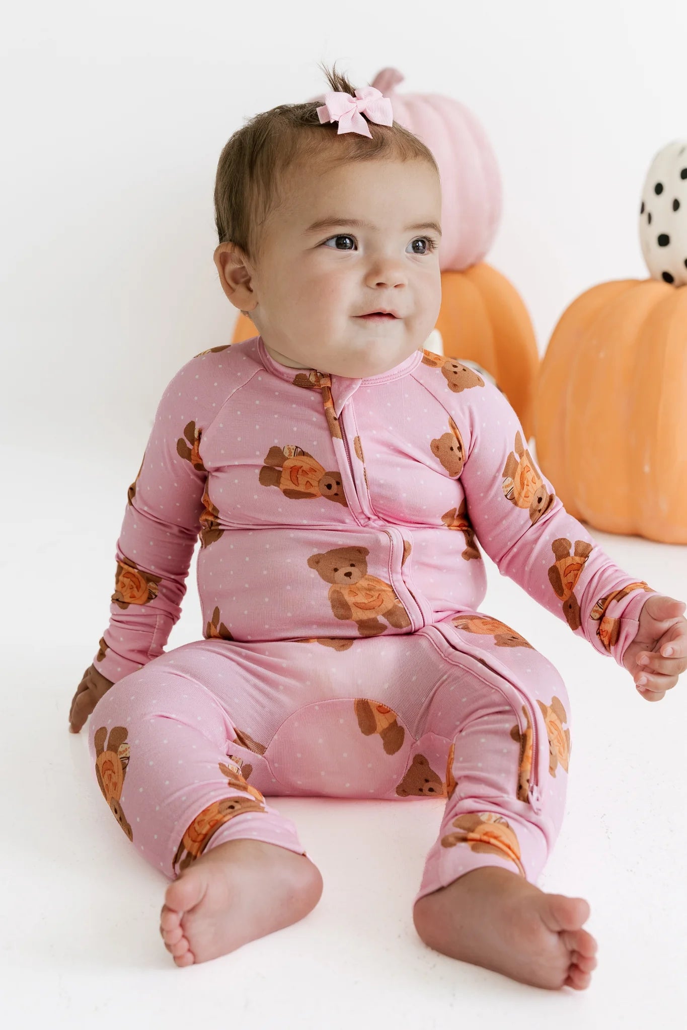 A baby wearing a pink long-sleeve zip-up pajama with a brown teddy bear print, sitting on a white studio floor with decorative pumpkins in the background.