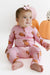 A baby wearing a pink long-sleeve zip-up pajama with a brown teddy bear print, sitting on a white studio floor with decorative pumpkins in the background.