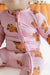 A close-up of a baby wearing a pink long-sleeve footless one-piece pajama with a front zipper and an all-over teddy bear print, seated against a soft orange and white background.
