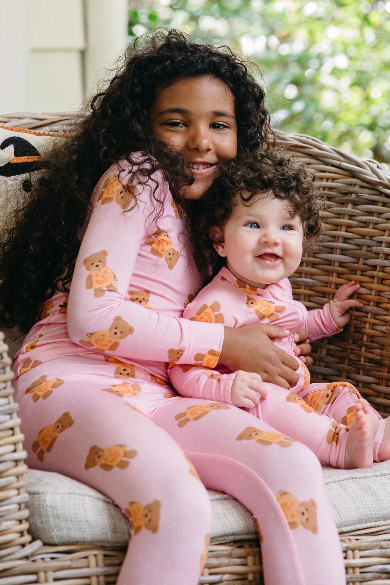 A smiling child and baby wearing matching pink long-sleeve pajamas with a brown teddy bear print, sitting together in a wicker chair.
