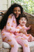 A smiling child and baby wearing matching pink long-sleeve pajamas with a brown teddy bear print, sitting together in a wicker chair.