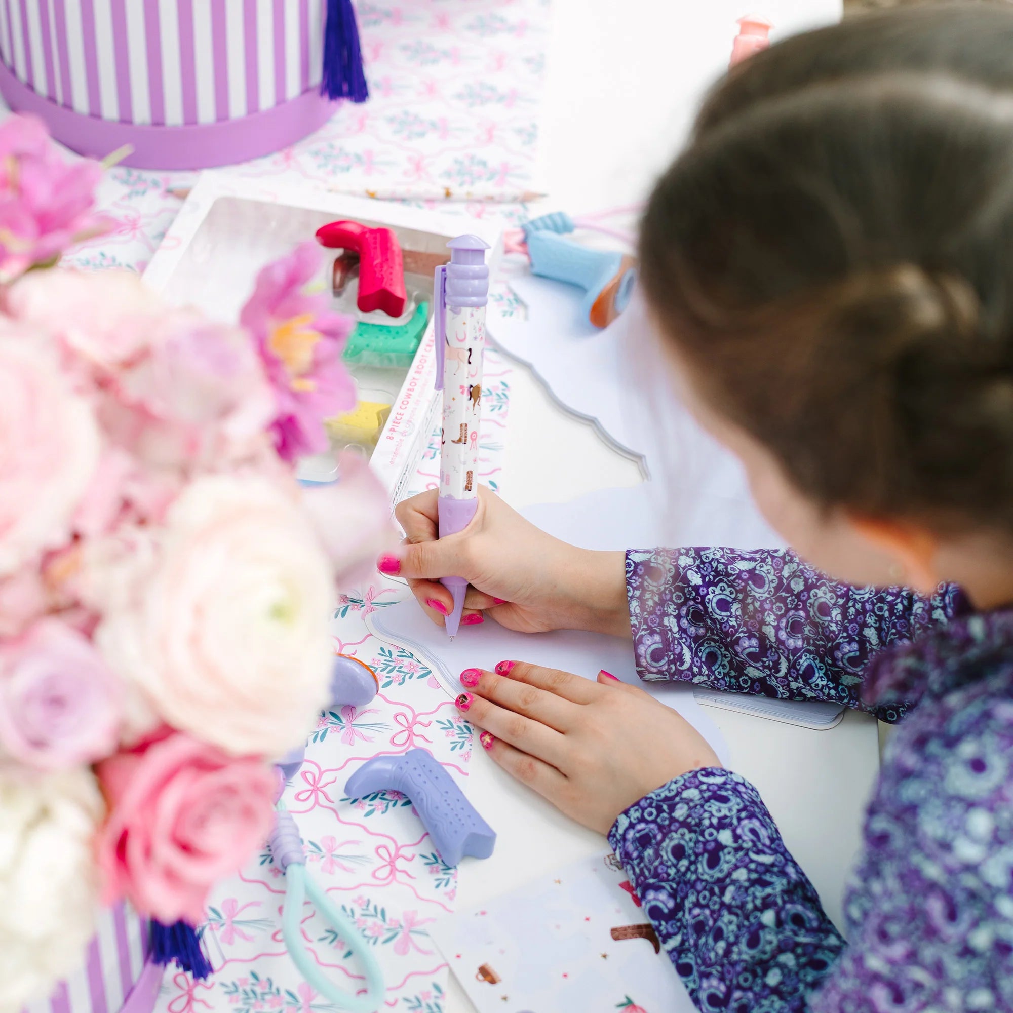 A child's hands and head are visible as they craft with a purple and white patterned pen or marker on a decorated table with flowers and craft supplies.