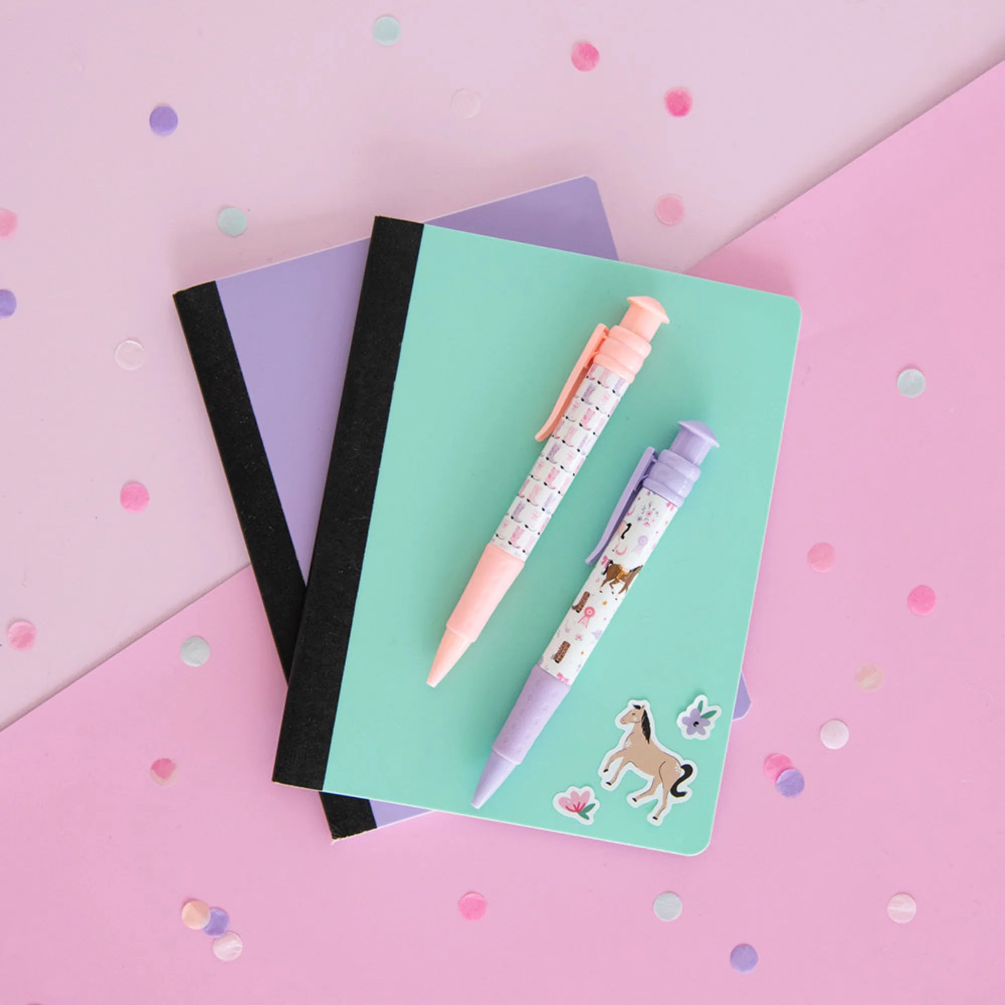 A styled flat-lay of pastel stationery on a pink background with confetti. The set includes a mint green notebook with a black elastic band, a lavender notebook underneath, a pink patterned pen, a lavender patterned pen, and small unicorn stickers.