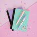 A styled flat-lay of pastel stationery on a pink background with confetti. The set includes a mint green notebook with a black elastic band, a lavender notebook underneath, a pink patterned pen, a lavender patterned pen, and small unicorn stickers.