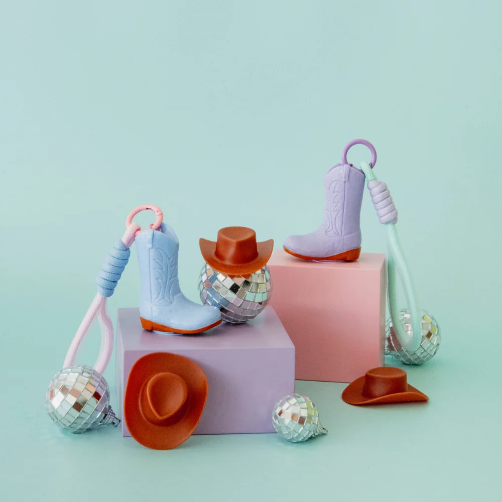 A set of pastel-colored toy cowboy boots, cowboy hats, and disco ball keychains arranged on pink display blocks against a light mint green background.