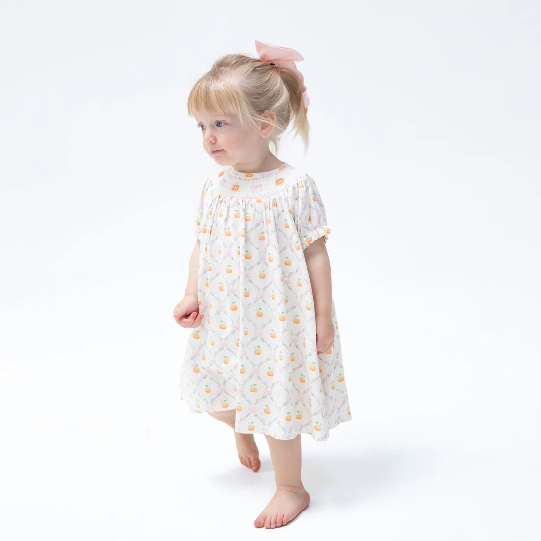 A young girl with blonde hair in a ponytail wearing a short-sleeve white smocked dress with a yellow and orange floral pattern, standing against a plain white background.