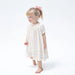 A young girl with blonde hair in a ponytail wearing a short-sleeve white smocked dress with a yellow and orange floral pattern, standing against a plain white background.