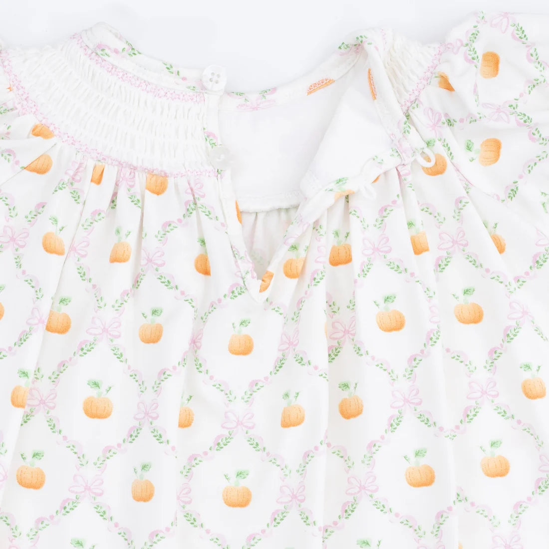 A close-up of a white baby dress featuring a repeating pattern of orange pumpkins, green vines, and pink bows, with a smocked neckline.
