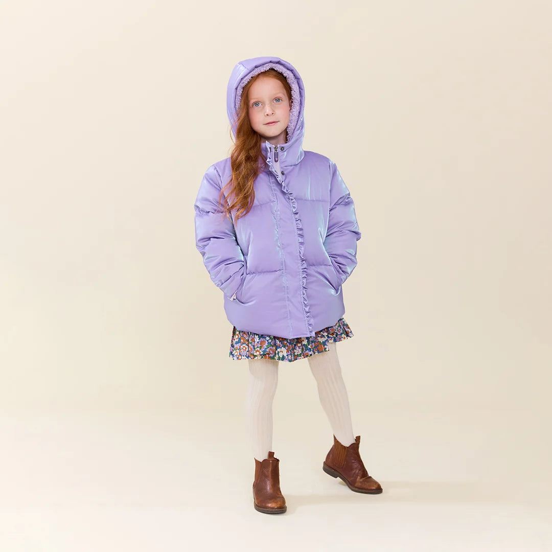 A young girl with long light brown hair stands against a solid beige studio background wearing a lavender purple hooded puffer jacket, a short floral print skirt, cream ribbed tights, and brown ankle boots.
