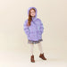 A young girl with long light brown hair stands against a solid beige studio background wearing a lavender purple hooded puffer jacket, a short floral print skirt, cream ribbed tights, and brown ankle boots.
