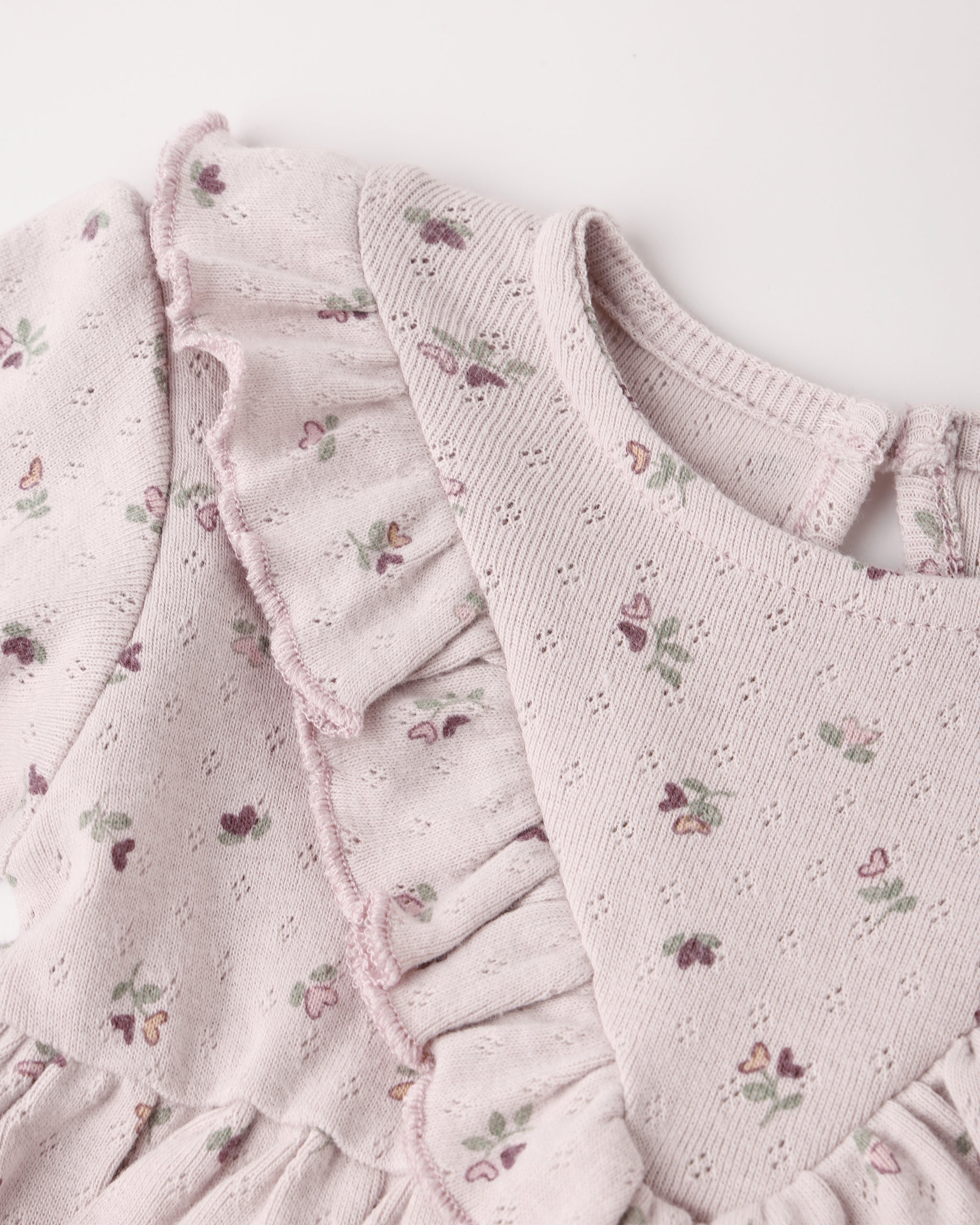 Close-up of a light lilac eyelet fabric garment with ruffled trim and a small floral print, showing the neckline and textured material against a plain light background.