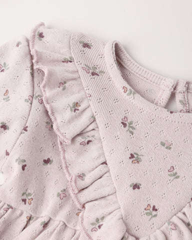 Close-up of a light lilac eyelet fabric garment with ruffled trim and a small floral print, showing the neckline and textured material against a plain light background.
