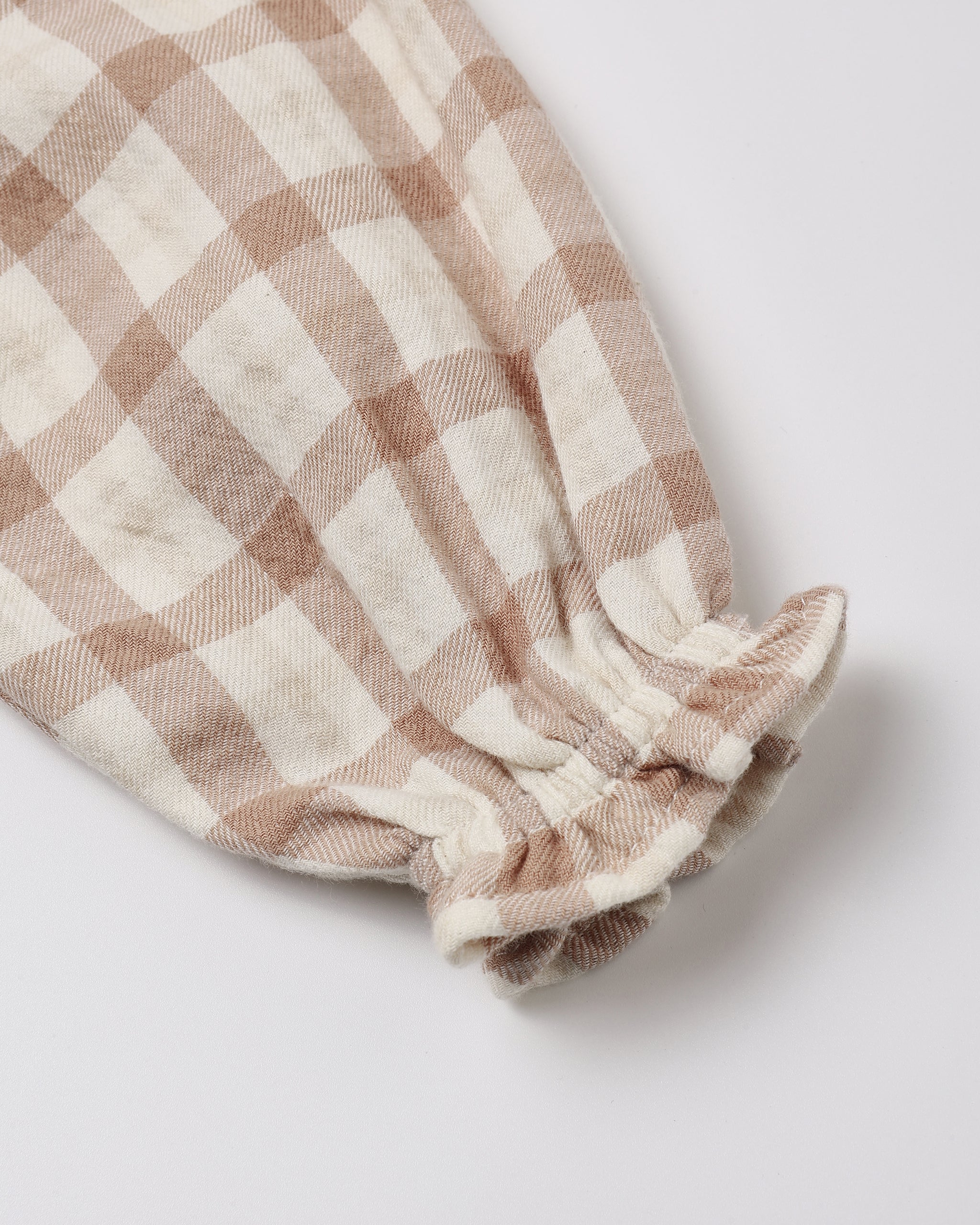 A close-up of a soft cotton fabric sleeve or cuff with a beige and off-white gingham check pattern and a gathered elastic ruffle at the end, shown on a light gray background.