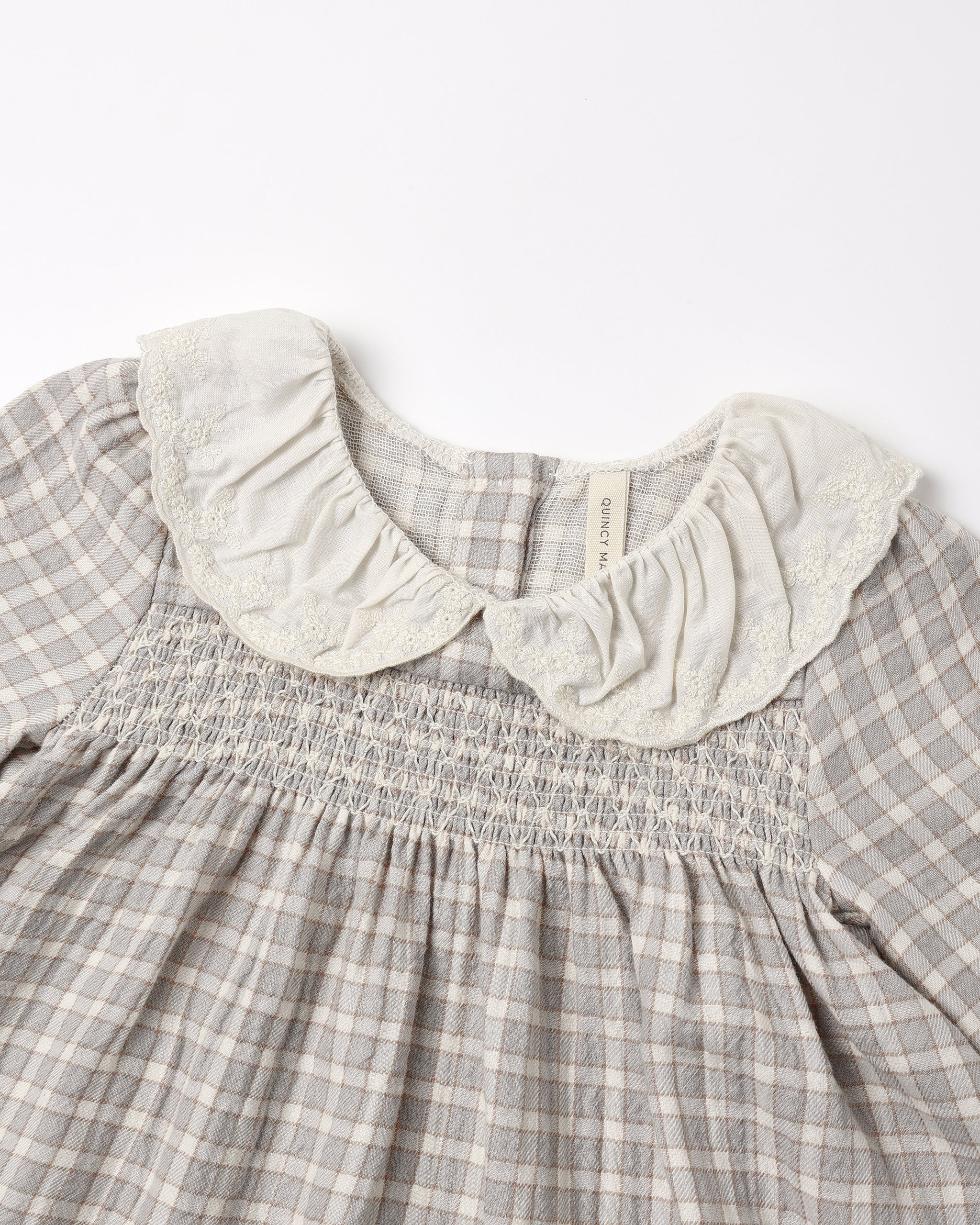 Close-up of a children's dress with a gray and white gingham pattern, featuring a smocked bodice and a large cream-colored ruffled lace Peter Pan collar.
