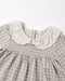 Close-up of a children's dress with a gray and white gingham pattern, featuring a smocked bodice and a large cream-colored ruffled lace Peter Pan collar.