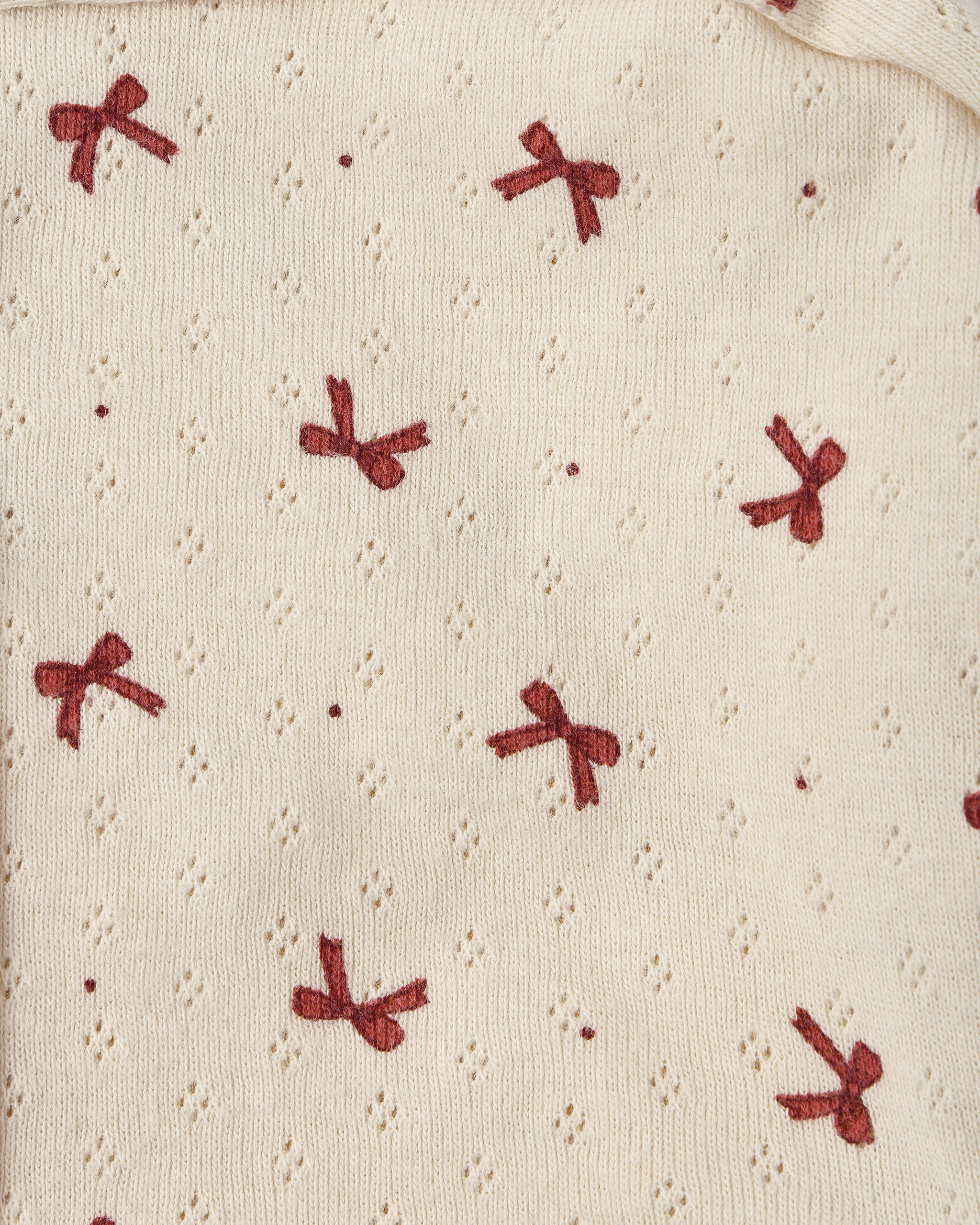 Close-up of a cream-colored knit fabric with a textured eyelet pattern and a repeating motif of small red embroidered bows.