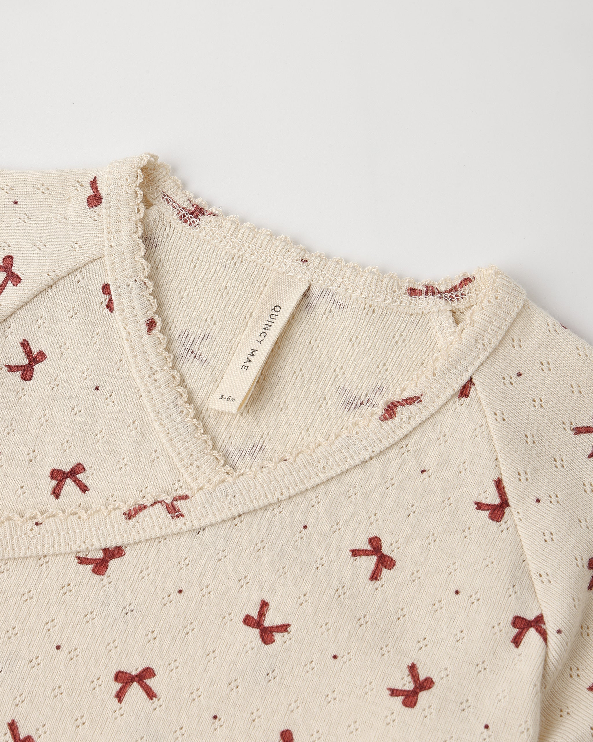 Close-up of a cream-colored knit top with a small red bow pattern and a visible 'Ouincy Mae' brand tag inside the neckline.
