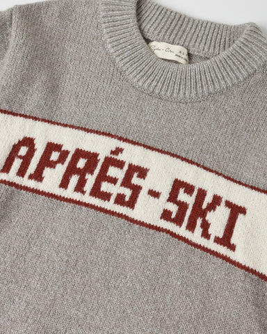 Close-up of a gray knit sweater with a cream horizontal stripe across the chest featuring the text 'APRÉS-SKI' in dark red.