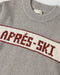 Close-up of a gray knit sweater with a cream horizontal stripe across the chest featuring the text 'APRÉS-SKI' in dark red.