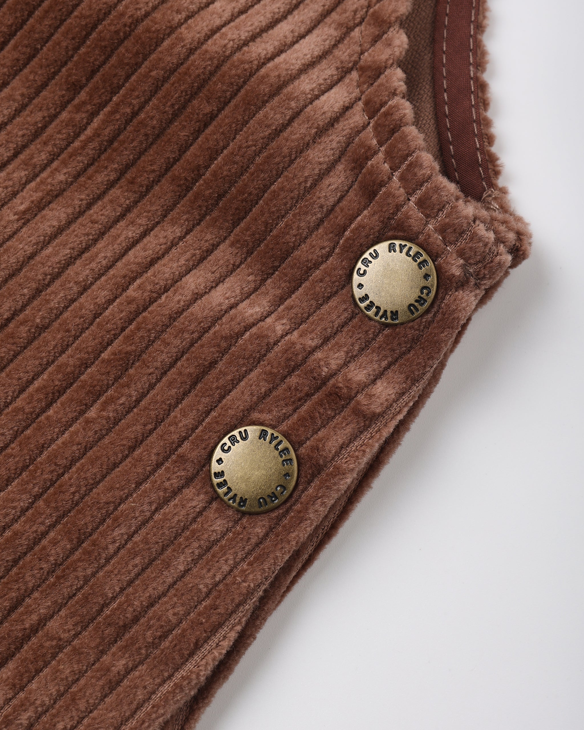 Close-up of a brown corduroy garment corner with two round brass buttons engraved with 'Cru Ayley', on a light gray background.