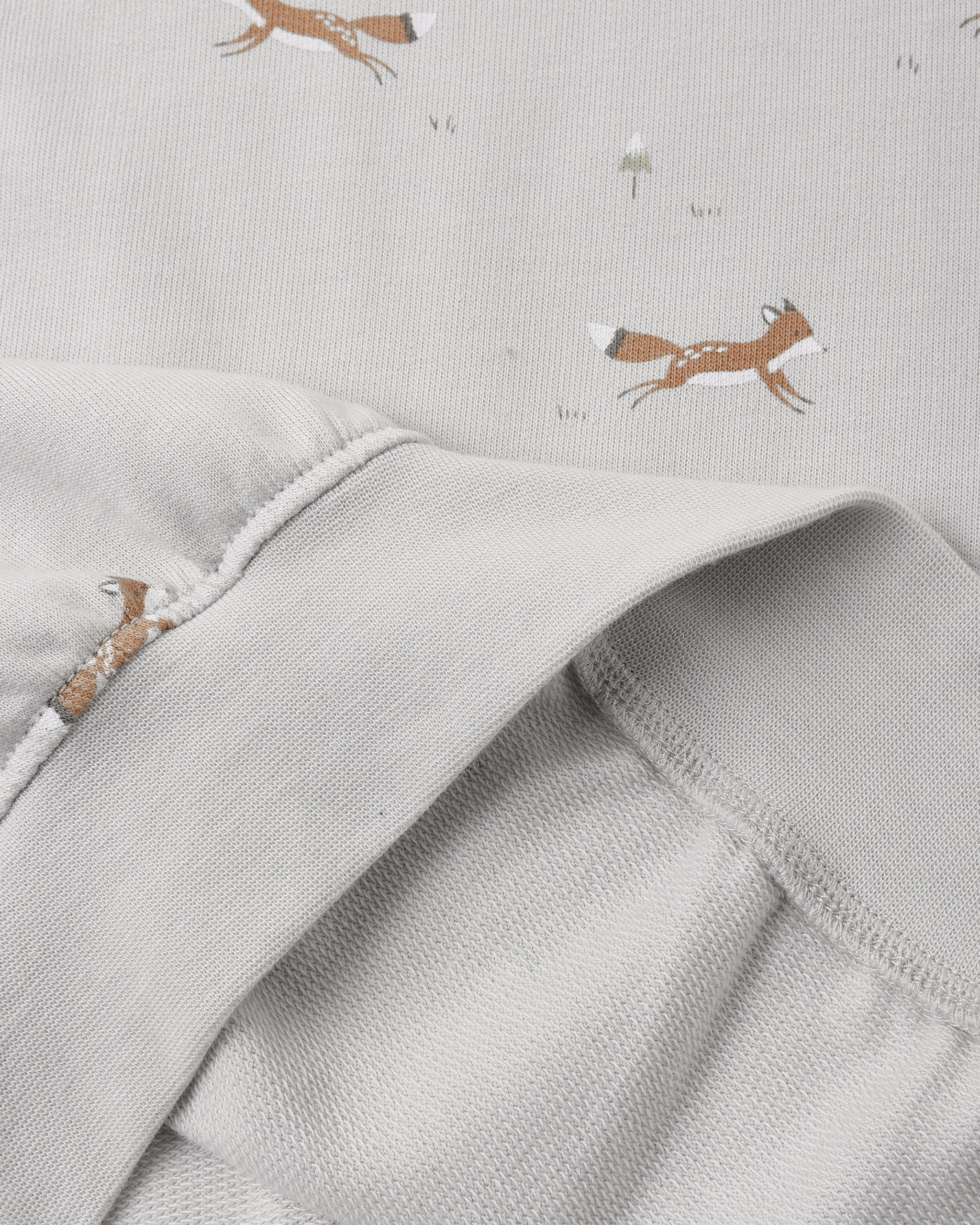 Close-up of a light gray fabric garment with a soft fold, showing a subtle print of brown foxes and small white dandelions on the fabric.