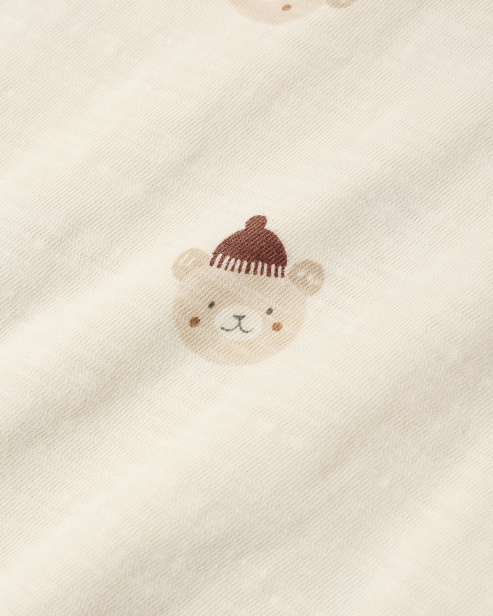 Close-up of a cream-colored fabric with a small embroidered teddy bear face wearing a brown knit hat.