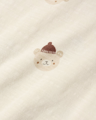 Close-up of a cream-colored fabric with a small embroidered teddy bear face wearing a brown knit hat.