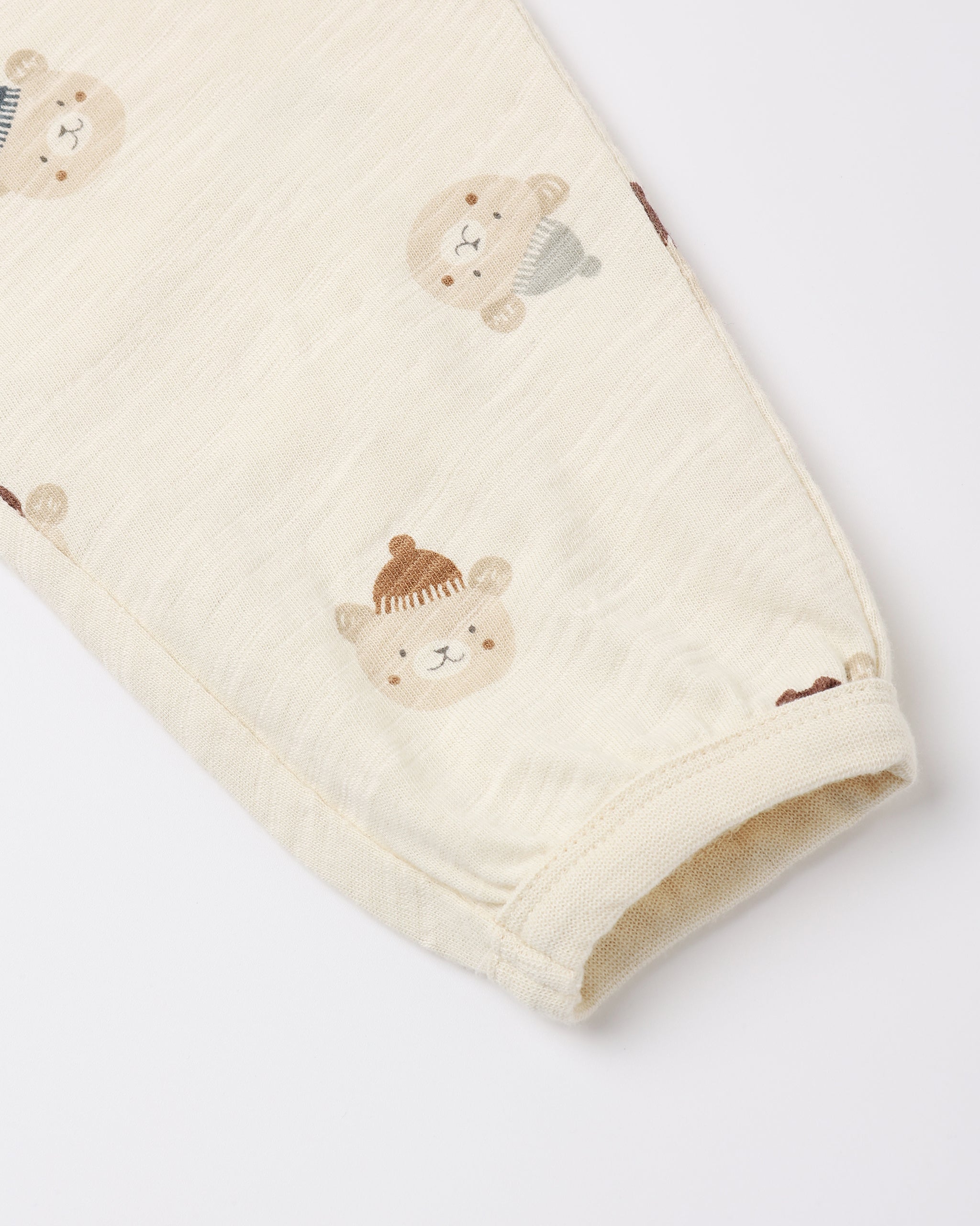 Close-up of a cream-colored baby garment, likely a sleeve or leg, featuring a soft fabric texture and an all-over print of teddy bear faces wearing knit hats.