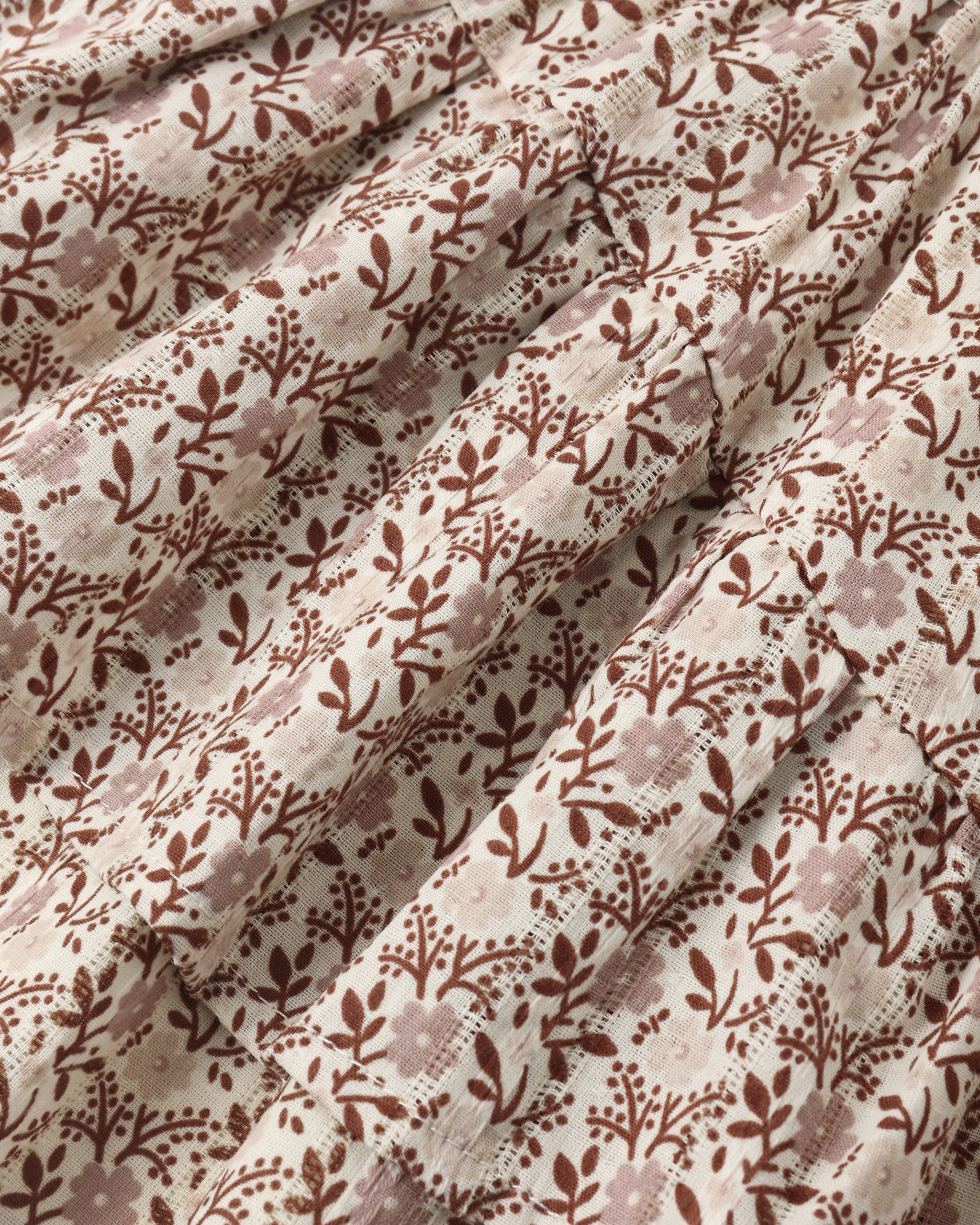 A close-up of a draped fabric with an all-over floral and leafy pattern in brown and dusty mauve on a warm off-white background.