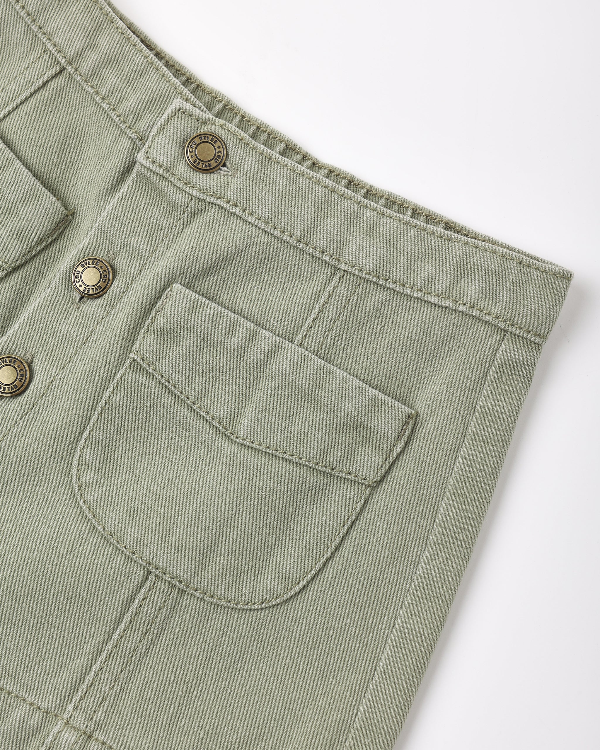Close-up of a light sage green corduroy garment, showing a buttoned pocket and metal buttons on a white background.
