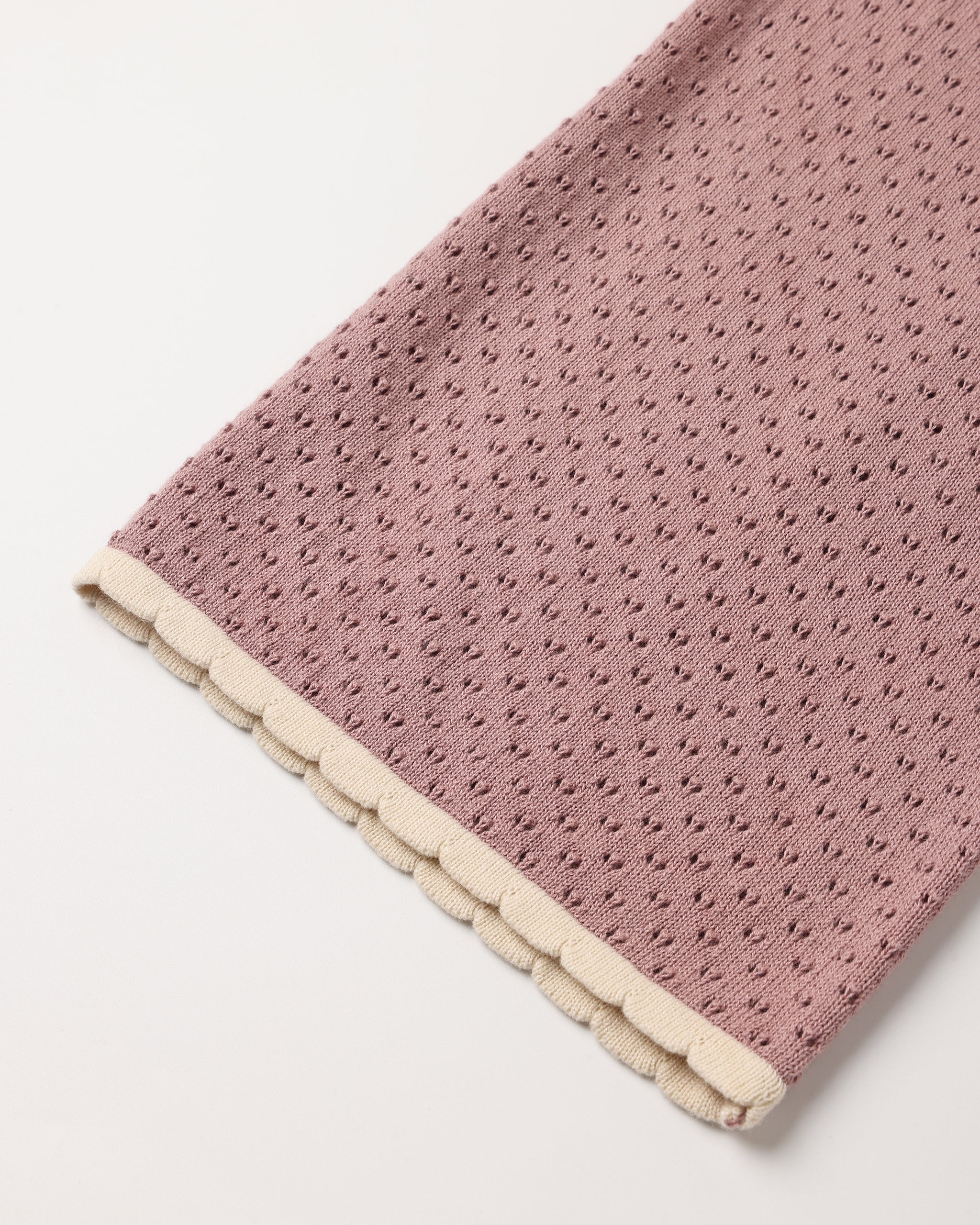 A close-up of a dusty pink textured fabric item, likely a blanket or scarf, featuring a scalloped cream border on a plain white background.