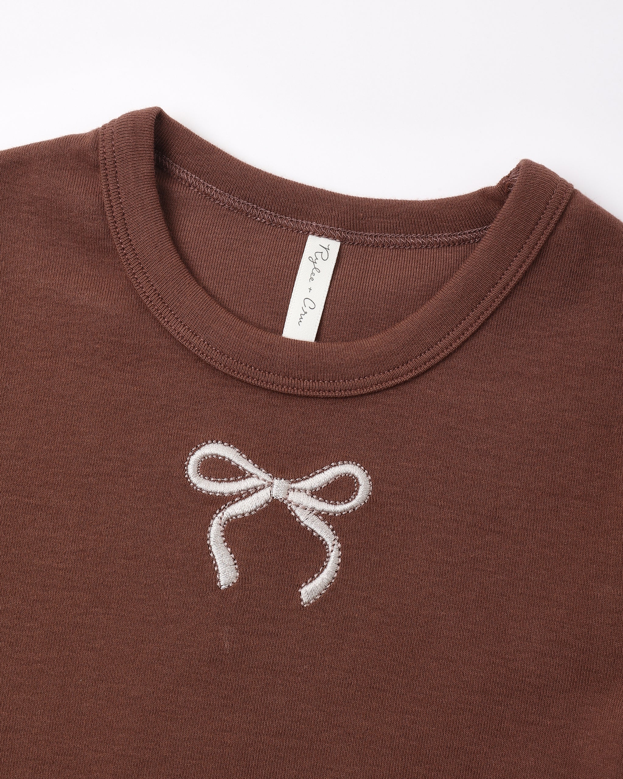Close-up of a brown crewneck t-shirt featuring a white embroidered bow on the chest, with a visible 'Pine Valley' brand tag inside the collar.