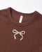 Close-up of a brown crewneck t-shirt featuring a white embroidered bow on the chest, with a visible 'Pine Valley' brand tag inside the collar.