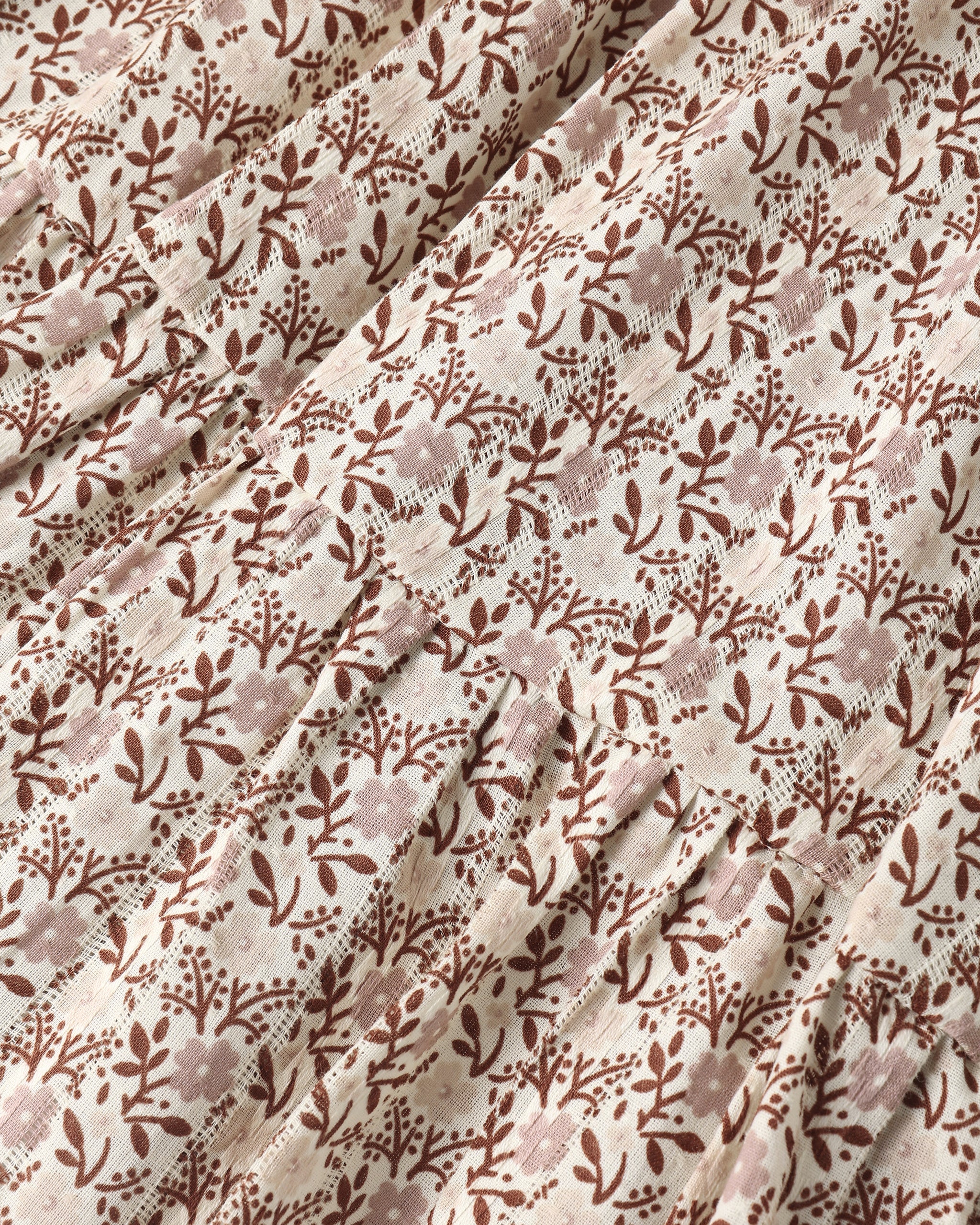 A close-up of a draped fabric with a repeating brown floral and leaf pattern on a warm cream background.