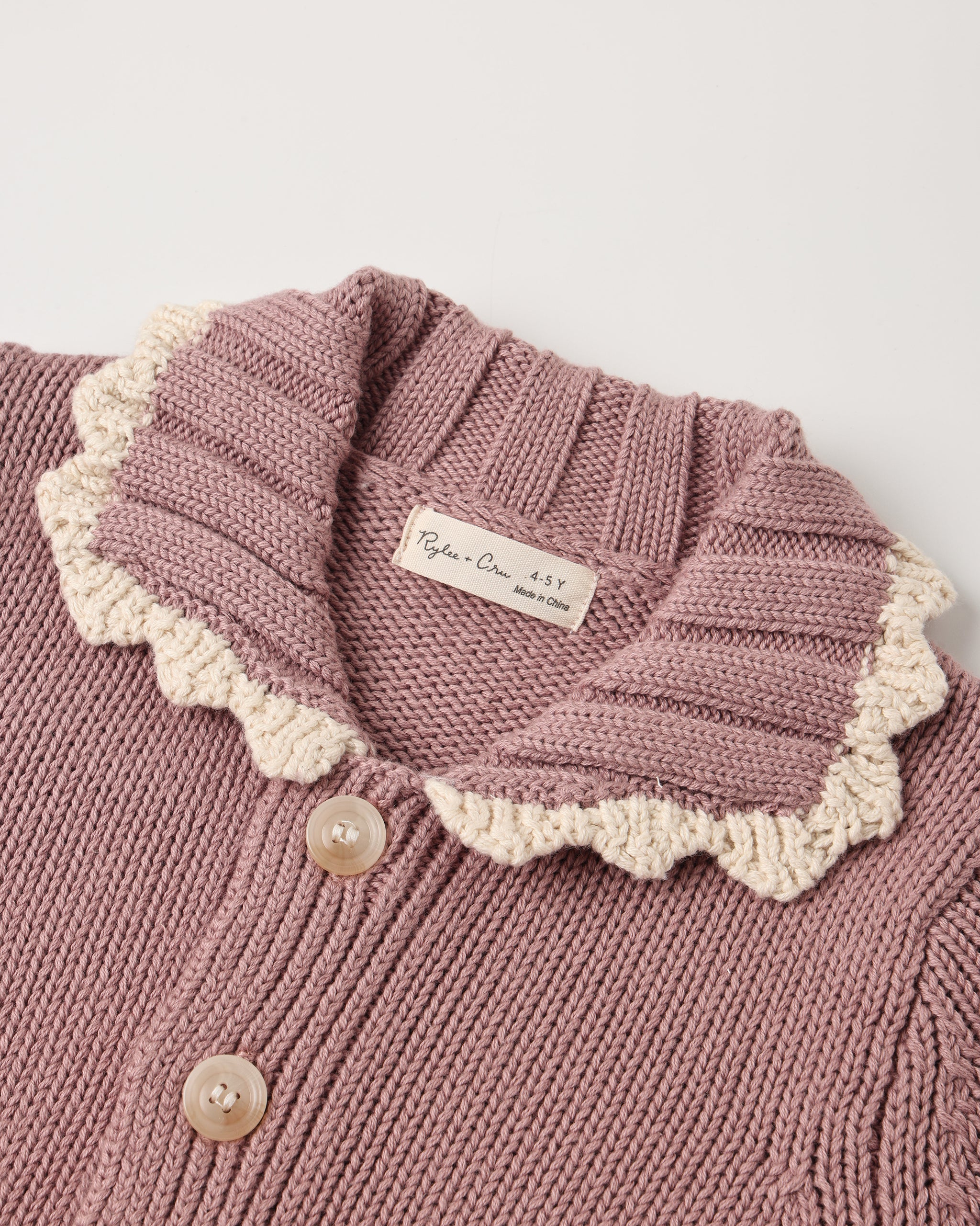 Close-up of a knitted cardigan in dusty mauve pink with a cream scalloped collar and light buttons, showing a brand label that reads 'Ryōn's Cream Sweaters'.