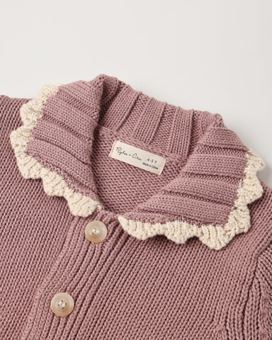 Close-up of a knitted cardigan in dusty mauve pink with a cream scalloped collar and light buttons, showing a brand label that reads 'Ryōn's Cream Sweaters'.