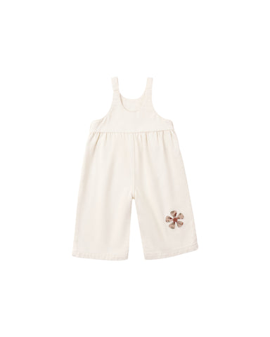 A natural colored denim overall with daisy patches applique shown  on a white background.