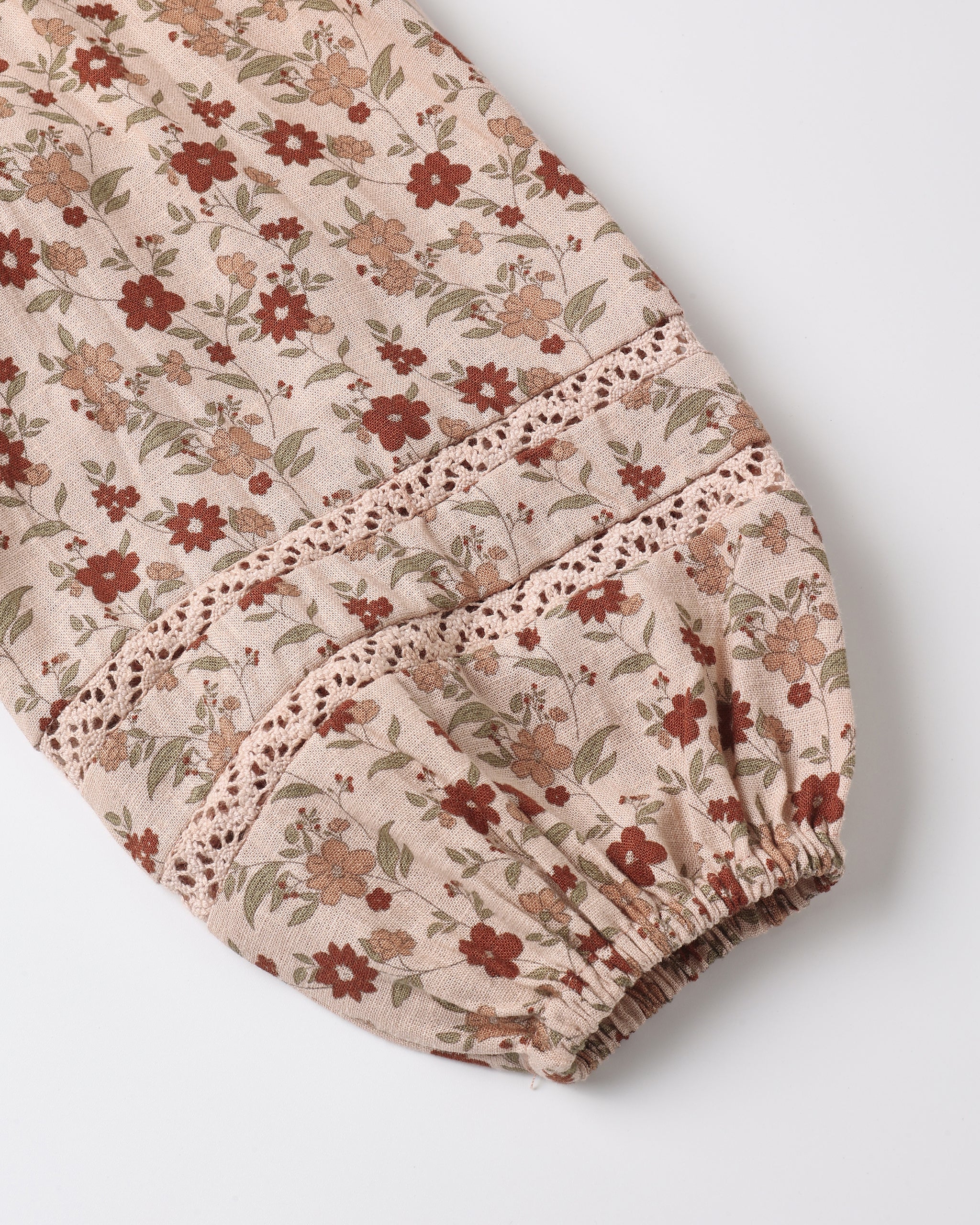A close-up of a floral-patterned fabric sleeve or garment section with a beige base, brown and red flowers, green leaves, and lace trim, finished with an elasticized ruffled edge, photographed on a light gray background.