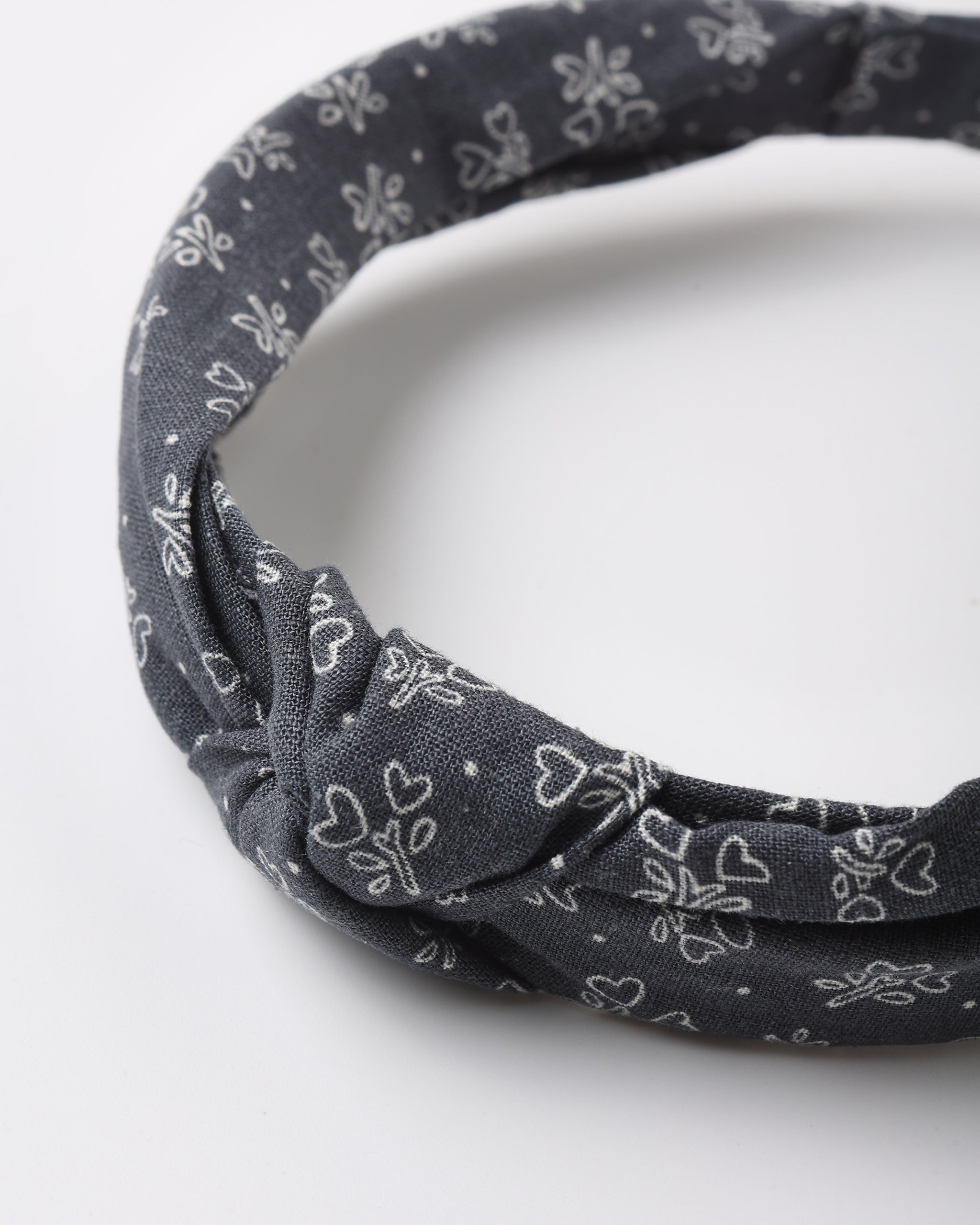 A dark gray fabric headband with a white floral and heart pattern, featuring a twisted knot detail at the front, photographed on a clean white background.