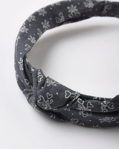 A dark gray fabric headband with a white floral and heart pattern, featuring a twisted knot detail at the front, photographed on a clean white background.