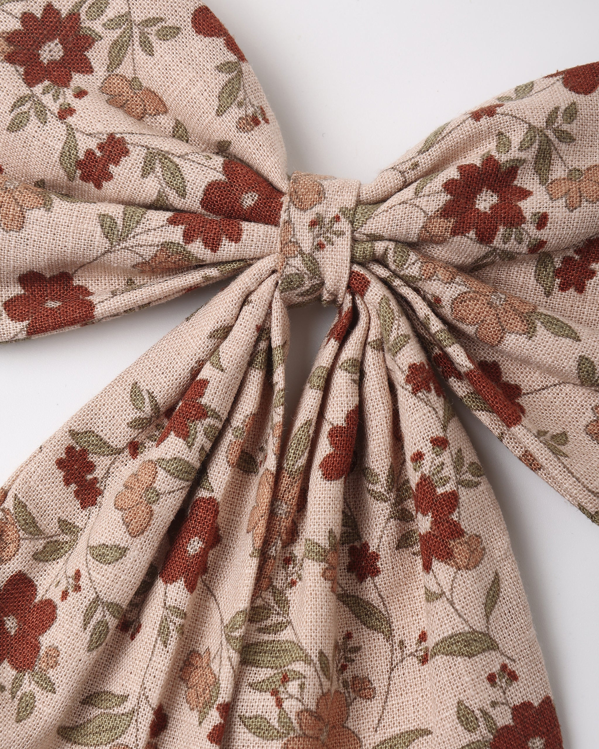 A close-up of a fabric hair bow with a floral pattern in rust red, olive green, and tan on a light beige background, tied in the center against a plain light gray backdrop.
