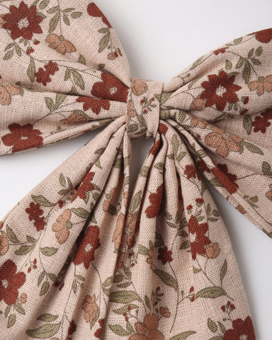 A close-up of a fabric hair bow with a floral pattern in rust red, olive green, and tan on a light beige background, tied in the center against a plain light gray backdrop.