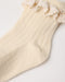 A close-up of a cream-colored ribbed knit sock with a matching lace ruffle trim at the top, laid flat on a light background.