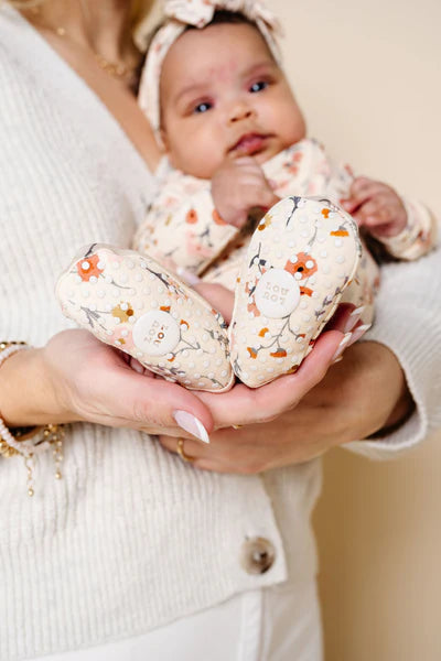 Baby holding a heart-shaped soft toy with floral patterns, wearing a white outfit and headband.