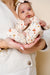 Baby holding a heart-shaped soft toy with floral patterns, wearing a white outfit and headband.