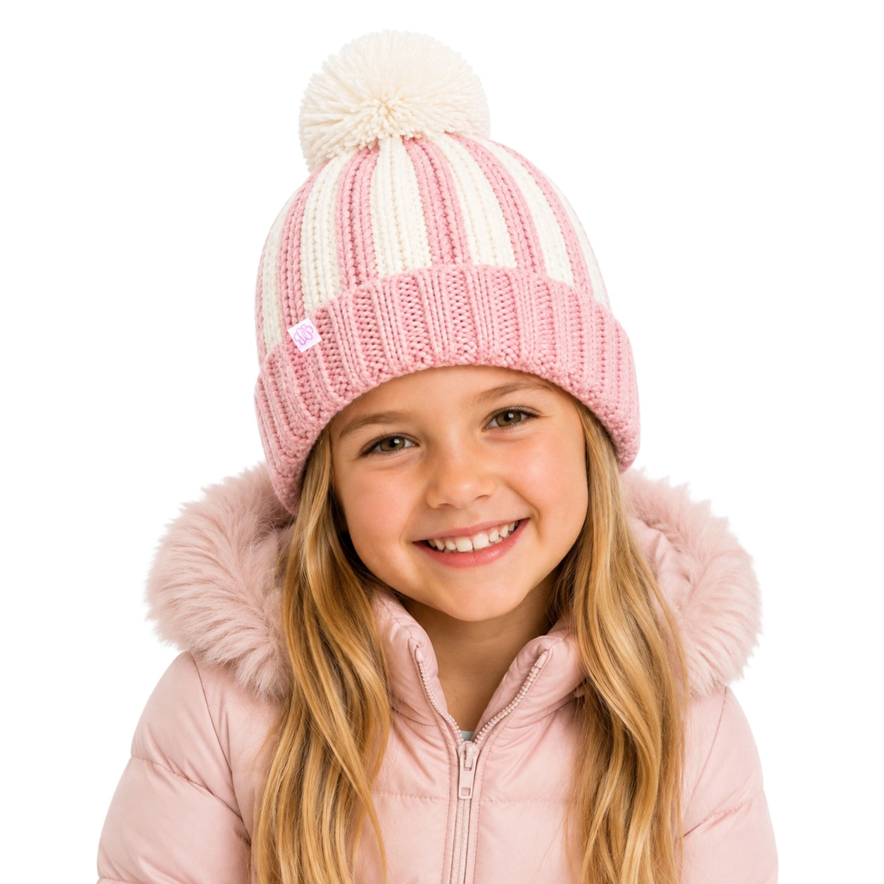 A young girl smiling while wearing a pink and cream striped knit beanie with a fluffy pom-pom, paired with a light pink hooded jacket against a solid white background.