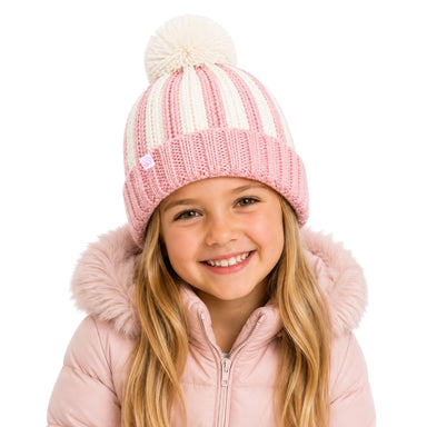 A young girl smiling while wearing a pink and cream striped knit beanie with a fluffy pom-pom, paired with a light pink hooded jacket against a solid white background.
