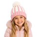 A young girl smiling while wearing a pink and cream striped knit beanie with a fluffy pom-pom, paired with a light pink hooded jacket against a solid white background.